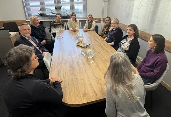 Jedenaście osób siedzi przy półokrągłym drewnianym stole w jasnym pokoju konferencyjnym z dużym oknem i roletami.