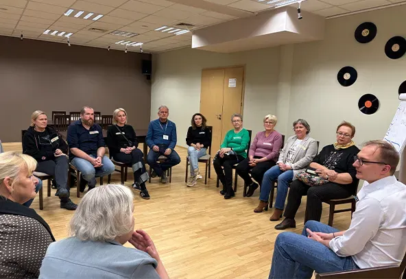 Grupa dwunastu osób siedzi na krzesłach w kręgu w jasnym pomieszczeniu z drewnianą podłogą. Ściana z tyłu jest jasna, wiszą na niej czarne płyty winylowe. Osoby rozmawiają lub słuchają, atmosfera wygląda na spotkanie lub warsztat.