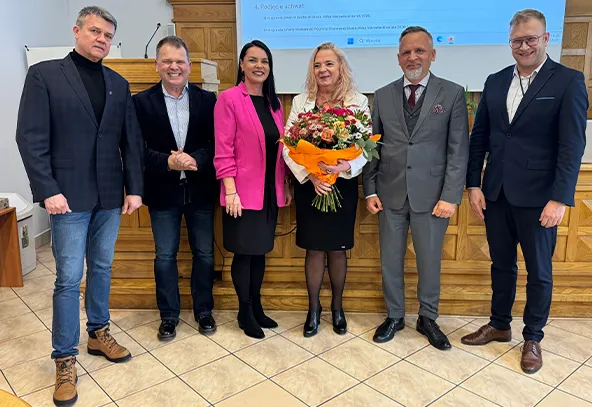 Sześć osób stoi w rzędzie przed drewnianą ścianą i podświetlanym ekranem. W środku kobieta w białej bluzce i czarnej spódnicy trzyma duży bukiet kwiatów owinięty w pomarańczowy papier. Po jej lewej stoi kobieta w różowym żakiecie, po obu stronach stoją mężczyźni w garniturach i marynarkach. Całość odbywa się w pomieszczeniu z jasnymi, kaflowymi płytkami na podłodze.