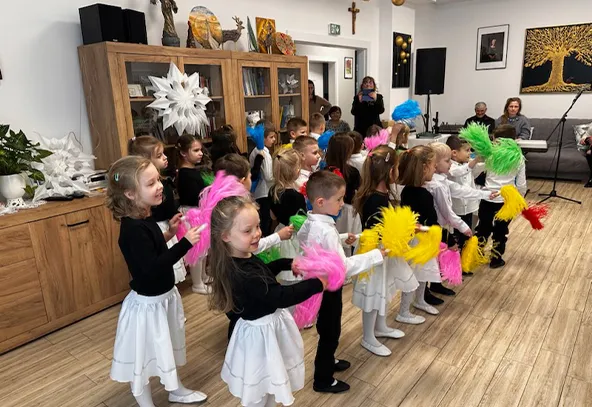 Około dwudziestu przedszkolaków stoi w rzędach w jasnej sali i macha kolorowymi pomponami (różowe, żółte, zielone, niebieskie). Dzieci noszą białe spódniczki lub spodnie oraz czarne lub białe góry. W tle drewniana szafka z białymi dekoracjami i kilka dorosłych obserwujących występ.