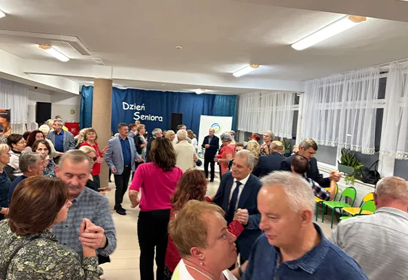 Wewnątrz sali wielu starszych i średniowiecznych ludzi tańczy i rozmawia. W tle niebieska zasłona z napisem "Dzień Seniora". Pomieszczenie jest jasno oświetlone, z białymi ścianami i dużymi oknami z firankami. Po prawej stronie stoją zielone krzesła i rośliny doniczkowe.