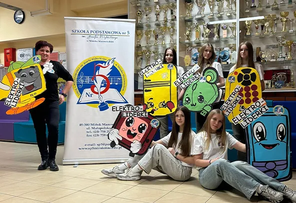 Siedem osób stoi i siedzi w holu szkolnym. Po lewej stoi dorosła kobieta, obok stoi pionowy baner z logo szkoły. W tle wysokie gabloty z licznymi pucharami. Kilka dziewcząt trzyma duże kolorowe plansze w kształcie rysunkowych postaci i urządzeń, dwie dziewczyny siedzą na podłodze z przodu.