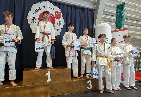 Siedmiu młodych zawodników w białych judogach stoi na podium i obok niego w hali sportowej. Trzech stoi na podium na miejscach 1 i 3, pozostali obok. Za nimi ciemny materiał z logo Judo Team. Każdy trzyma dyplom, niektórzy mają medale.
