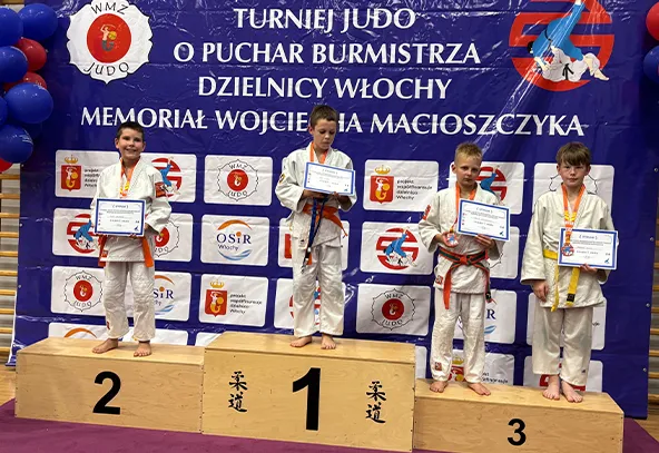 Czterech chłopców w judogach stoi na podium podczas turnieju judo, każdy trzyma dyplom i medal, za nimi niebieski baner z nazwą turnieju i sponsorami.