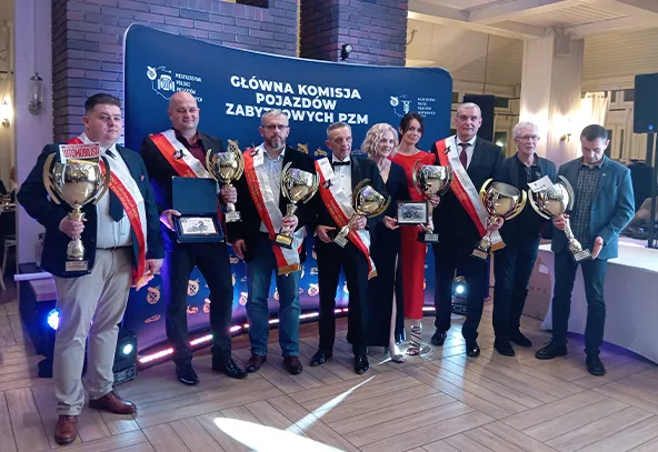 Osiem osób stoi w rzędzie na tle niebieskiego baneru z napisem dotyczącym głównej komisji pojazdów zabytkowych. Sześć osób trzyma puchary i tabliczki z nagrodami. Kilka osób nosi czerwono-białe szarfy. Wnętrze jasne, podłoga drewniana, oświetlenie sztuczne.