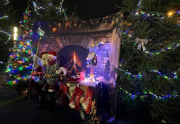 Kobieta siedzi obok osoby przebranej za Świętego Mikołaja na tle dekoracji kominka z płonącym ogniem. Wokół stoją udekorowane świątecznymi światłami choinki. Scena jest oświetlona kolorowymi lampkami w nocy.