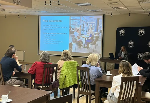W sali konferencyjnej około dziewięciu osób siedzi przy drewnianych stołach. Kobieta przy biurku obsługuje laptop i prowadzi prezentację na dużym ekranie. Ekran wyświetla slajd z tekstem i fotografią. Pomieszczenie jest jasno oświetlone, sufit z panelami i lampami. Na ścianie obok wisi ciemny panel z powtarzającym się białym logo.