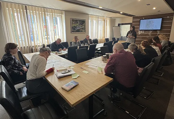 W sali konferencyjnej przy długim drewnianym stole siedzi dziesięć osób. Kilka osób zapisuje notatki. Jedna osoba stoi przy telewizorze z wyświetloną prezentacją. Przy oknach pionowe jasne żaluzje wpuszczające dzienne światło. Ściany jasne, na jednej wisi obraz.