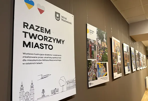 Wewnętrzna galeria z szarobrązową ścianą, przy której w rzędzie wiszą duże drukowane plakaty i fotografie. Po lewej duży biały plakat z dwoma kolorowymi sercami w górnym rogu i pogrubionym czarnym napisem, dalej kilka kolorowych plansz z fotografiami wydarzeń.