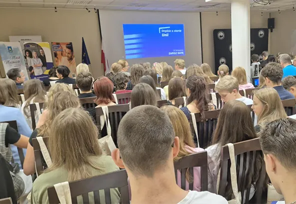 Wnętrze sali konferencyjnej z widoczną grupą około trzydziestu siedzących osób zwróconych przodem do ekranu projekcyjnego na ścianie. Ekran wyświetla prezentację na niebieskim tle. W tle po lewej stronie widoczne flagi oraz plakaty informacyjne.