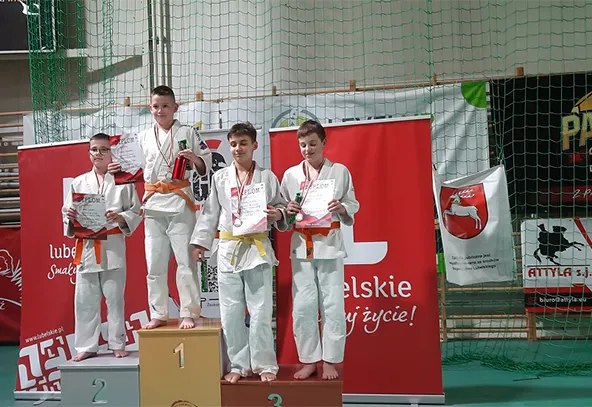 Czterech chłopców w białych judogach stoi na podium w sali sportowej, każdy trzyma dyplom i medal. Tło to siatka i czerwony baner z białymi napisami.