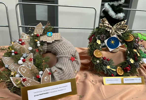 Dwa świąteczne wieńce stojące na beżowym materiale na stole. Wieniec po lewej stronie jest szary, ozdobiony kartonowymi domkami, czerwonymi i białymi kulkami oraz zielonymi gałązkami. Wieniec po prawej stronie jest zielony, udekorowany suszonymi plasterkami pomarańczy, czerwonymi jagodami, złotymi gwiazdkami i kokardą w kratę. Za wieńcami widać okno i metalową barierkę.