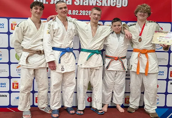 Pięciu młodych mężczyzn stoi obok siebie na tle czerwonego baneru i ściany z logo sponsorów. Wszyscy noszą białe kimona judo. Od lewej: pierwszy ma ciemne włosy i biały pas, drugi ma krótko ostrzyżone włosy i niebieski pas, trzeci ma zielony pas i rękę na ramieniu drugiego, czwarty ma pomarańczowo-zielony pas, piąty ma pomarańczowy pas, medal na szyi i trzyma dyplom. Wszystkie osoby stoją na czerwonej podłodze wewnątrz hali.