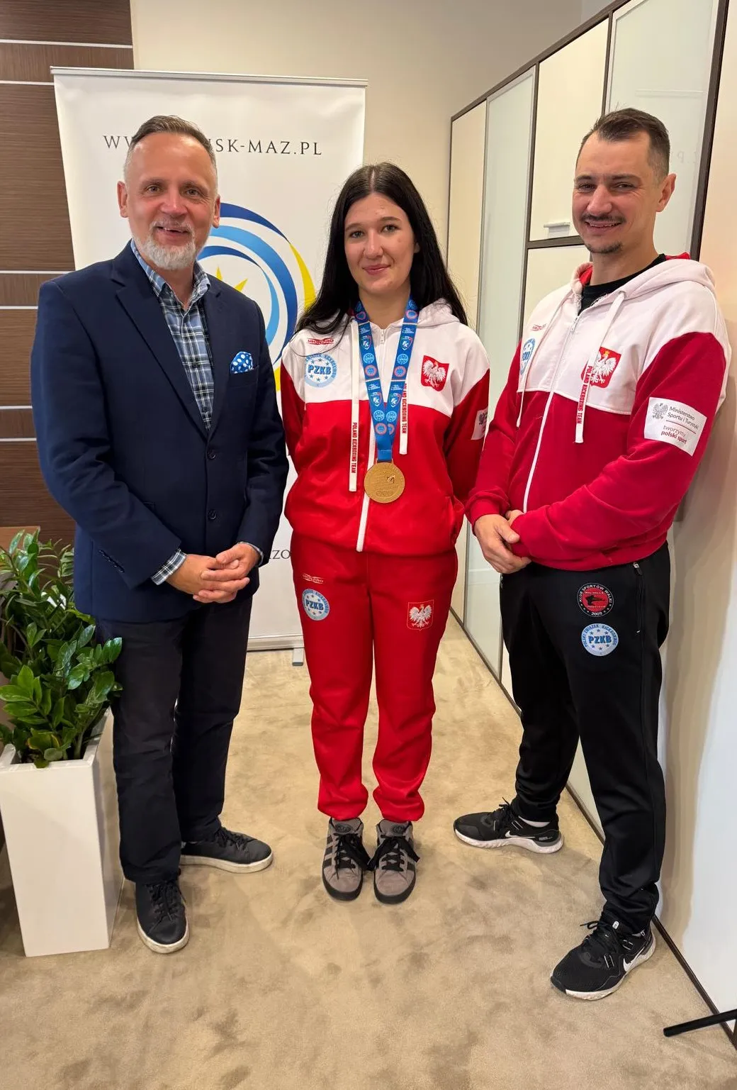 Trzy osoby stoją wewnątrz pomieszczenia na beżowym dywanie. Środkowa kobieta w czerwono-białym dresie z medalem na szyi. Po lewej mężczyzna w granatowej marynarce i ciemnych spodniach. Po prawej mężczyzna w czerwono-białym dresie i czarnych spodniach. W tle biała ściana i roślina doniczkowa.