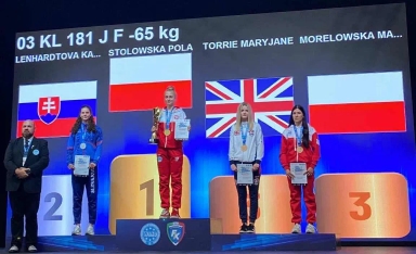 Cztery zawodniczki stoją na podium z medalami i dyplomami, w tle widoczne flagi Słowacji, Polski i Wielkiej Brytanii oraz oznaczenia kategorii wagowej -65 kg. Sędzia stoi obok podium.