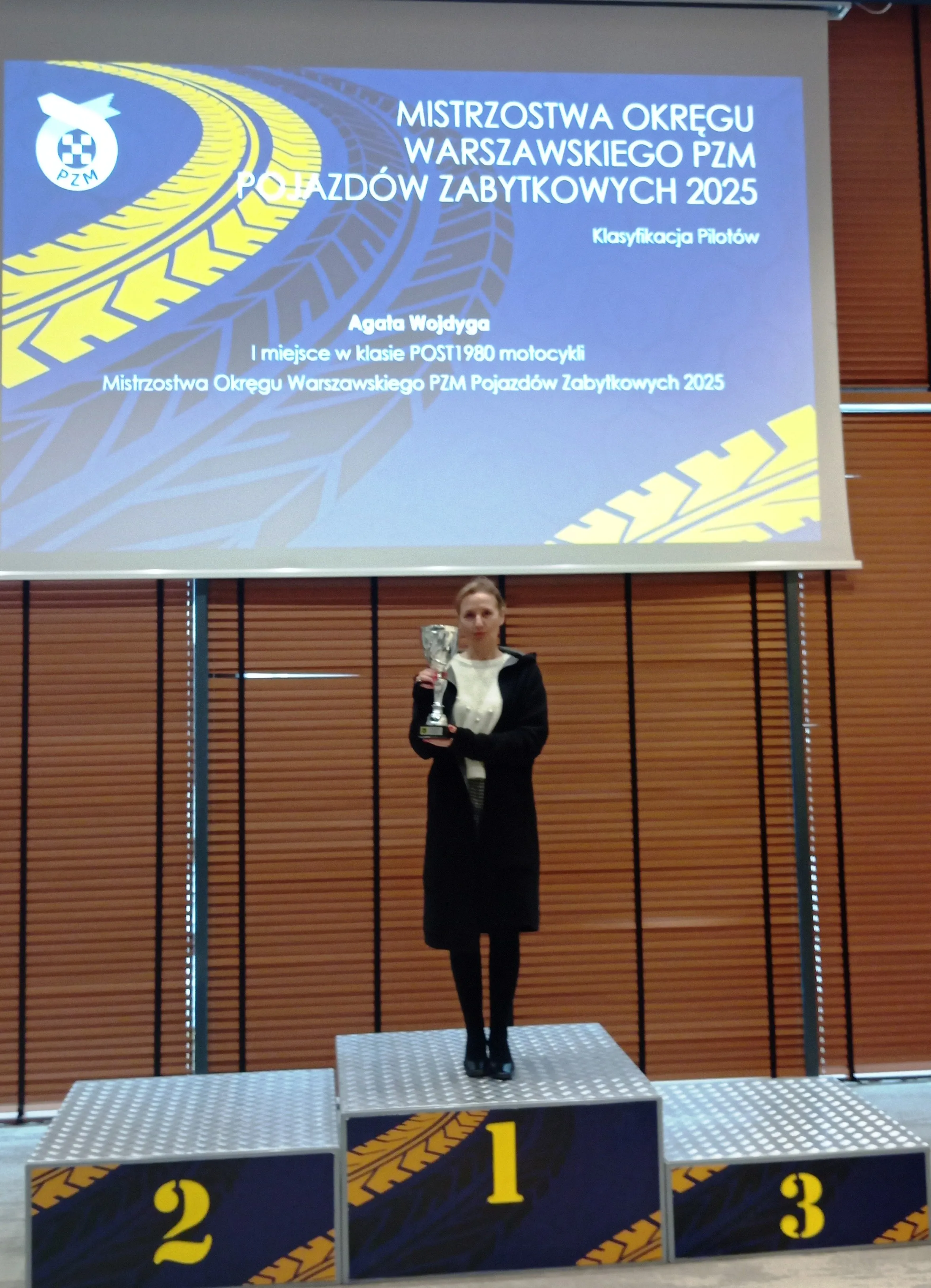 Kobieta stoi na podium oznaczonym numerem 1 i trzyma puchar. Sala wewnątrz z zasłoniętymi drewnianymi żaluzjami za nią. Nad podium wisi duży niebieski ekran z żółym wzorem opony. Kobieta ma ciemny płaszcz i jasną górę.