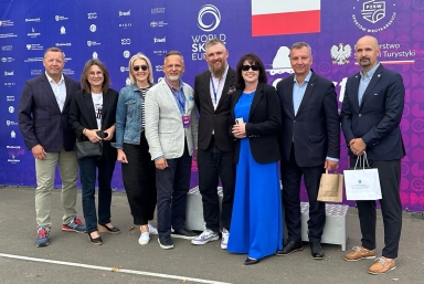 Grupa ośmiu osób stoi na zewnątrz na tle niebiesko-fioletowego baneru z logo World Skate Europe i flagą Polski, wszyscy ubrane formalnie lub półformalnie, część osób trzyma torby lub telefon.