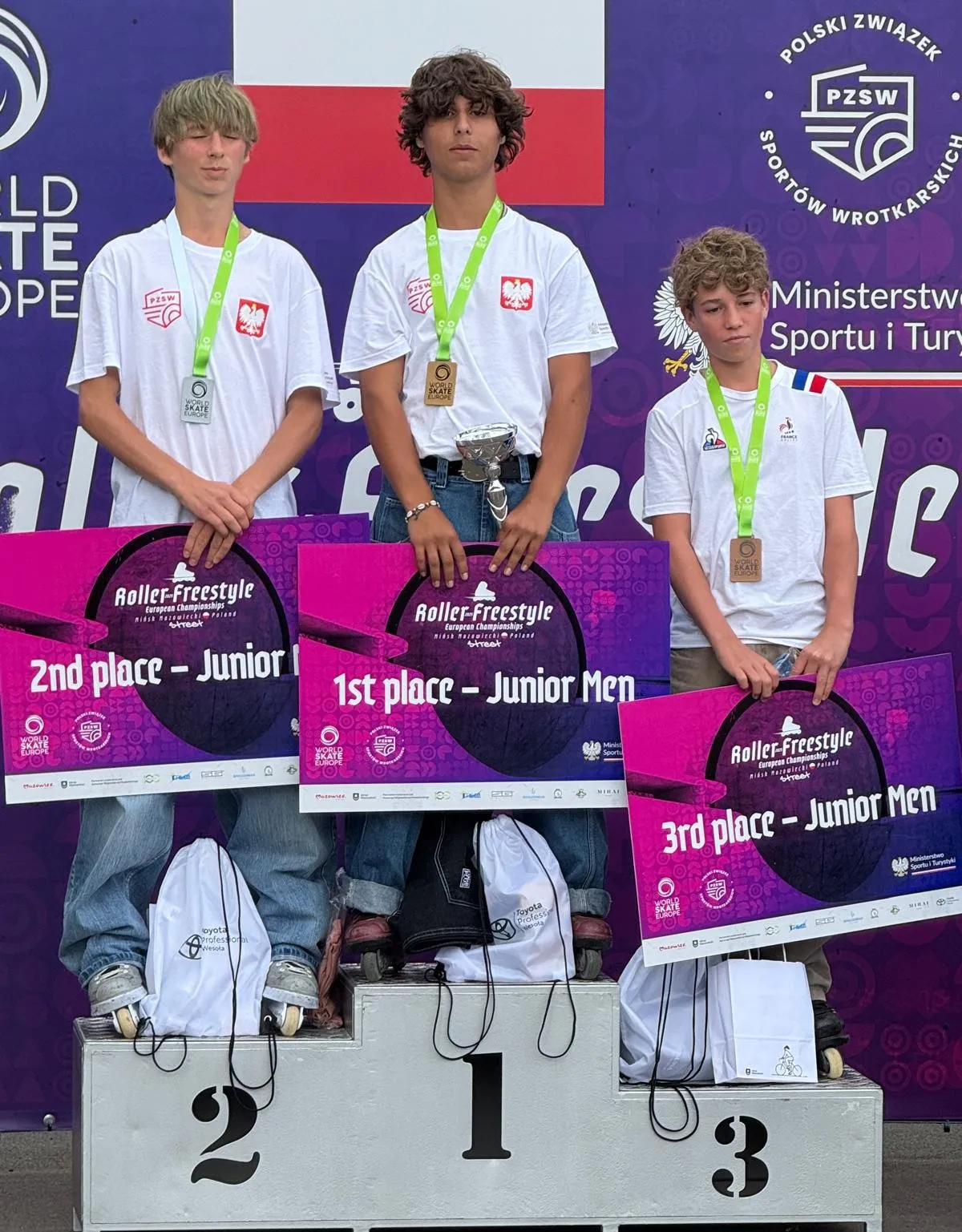 Trzech chłopców z medalami na szyi stoi na podium z miejscami 1., 2. i 3., trzymając tablice informujące o kolejności zajętych miejsc w kategorii Junior Men w zawodach Roller Freestyle.