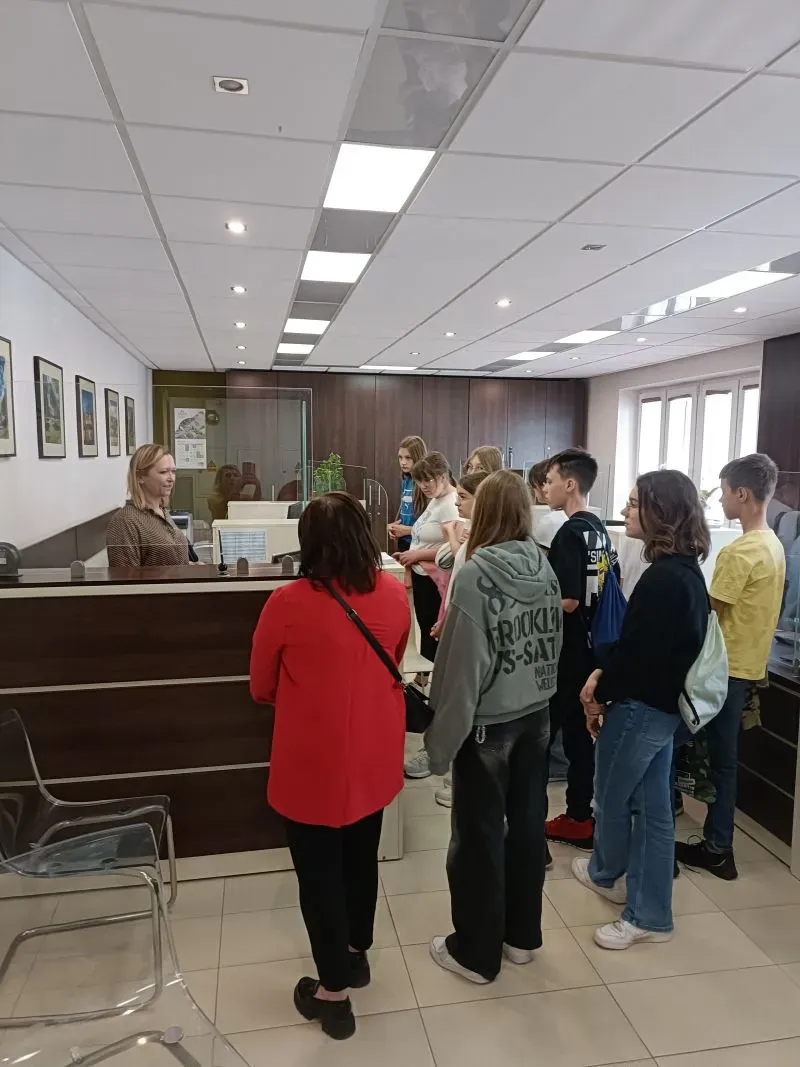 Grupa młodych osób stoi przy ladzie rozmawiając z siedzącą po drugiej stronie kobietą.