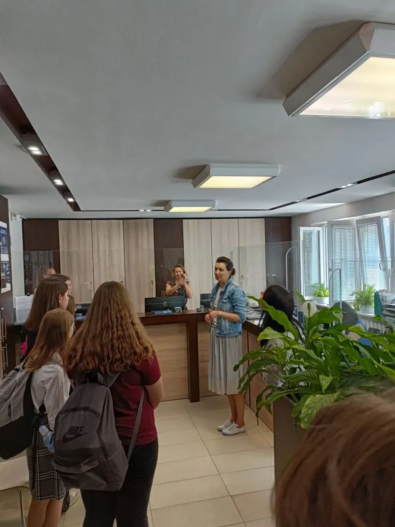 Grupa osób stoi w biurze przed recepcją, gdzie dwie kobiety rozmawiają, a wokół znajdują się rośliny i meble biurowe.