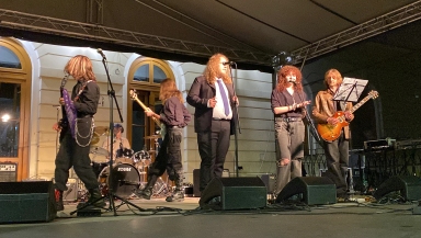 Sześć osób na scenie podczas koncertu na zewnątrz w nocy, trzech mężczyzn gra na gitarach, jedna osoba siedzi przy perkusji, dwie osoby stoją przy mikrofonach, budynek oświetlony w tle.