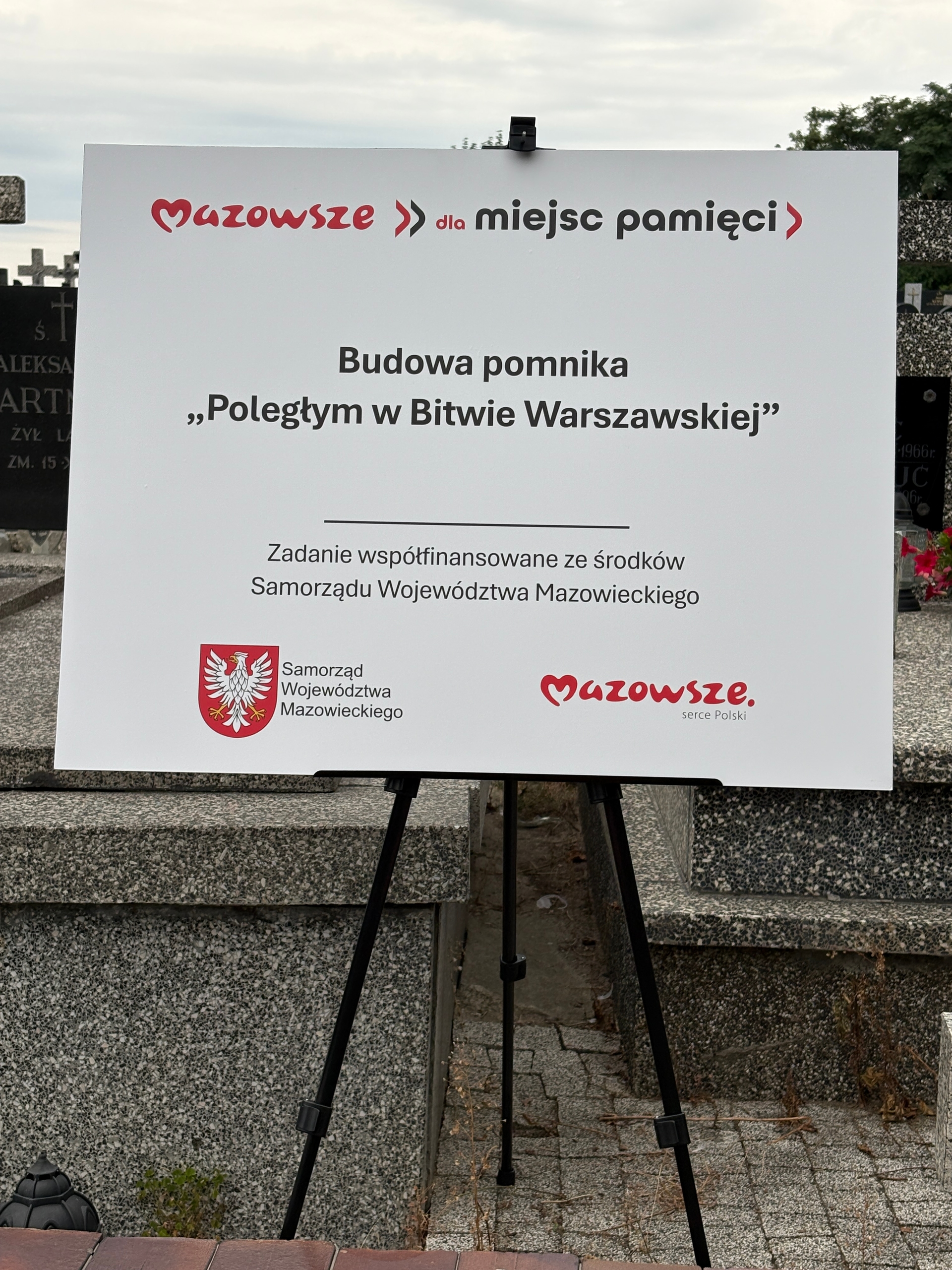 Biała tablica informacyjna na czarnym statywie stojąca na cmentarzu. Tablica ma czerwone logo i herb województwa w dolnej części. W tle widoczne nagrobki i drzewa. Dzień, niebo zachmurzone.
