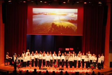 Grupa dzieci i nastolatków stoi na scenie teatralnej w rzędzie, ubrani na biało i czarno. Z lewej strony gra osoba przy fortepianie. W tle na dużym ekranie wyświetlany jest obraz krajobrazu z widokiem na poranne pola i drzewa oświetlone wschodzącym słońcem. Na pierwszym planie stoją kwiaty i świece. Publikum siedzi na widowni. Scena jest oświetlona ciepłym, czerwonym światłem.