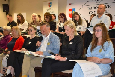 Grupa dorosłych i młodych osób siedzi na krzesłach w sali podczas wydarzenia związanego z czytaniem, niektórzy trzymają mikrofony i czytają z książek lub kartek.