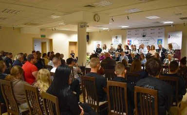 Grupa osób siedzi na krzesłach w sali podczas wydarzenia Narodowe Czytanie, część z nich czyta teksty na scenie z mikrofonem.