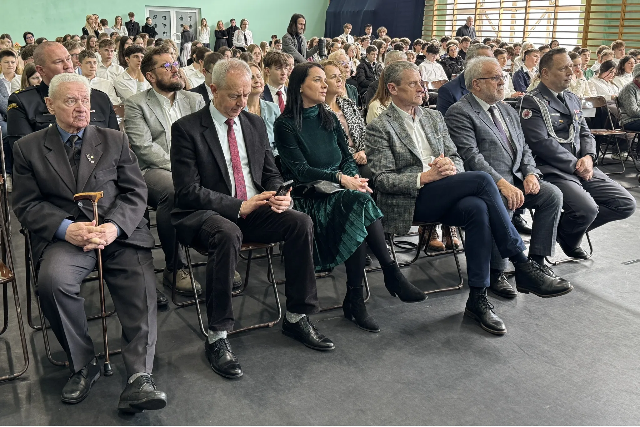 Grupa osób siedzi w rzędach krzeseł na sali gimnastycznej. W pierwszym rzędzie siedzi starszy mężczyzna z laską, mężczyzna w garniturze z telefonem, kobieta w zielonej sukience oraz trzej mężczyźni w marynarkach i mundurze. W tle siedzi wiele młodych osób w białych koszulach przy zielonej ścianie.