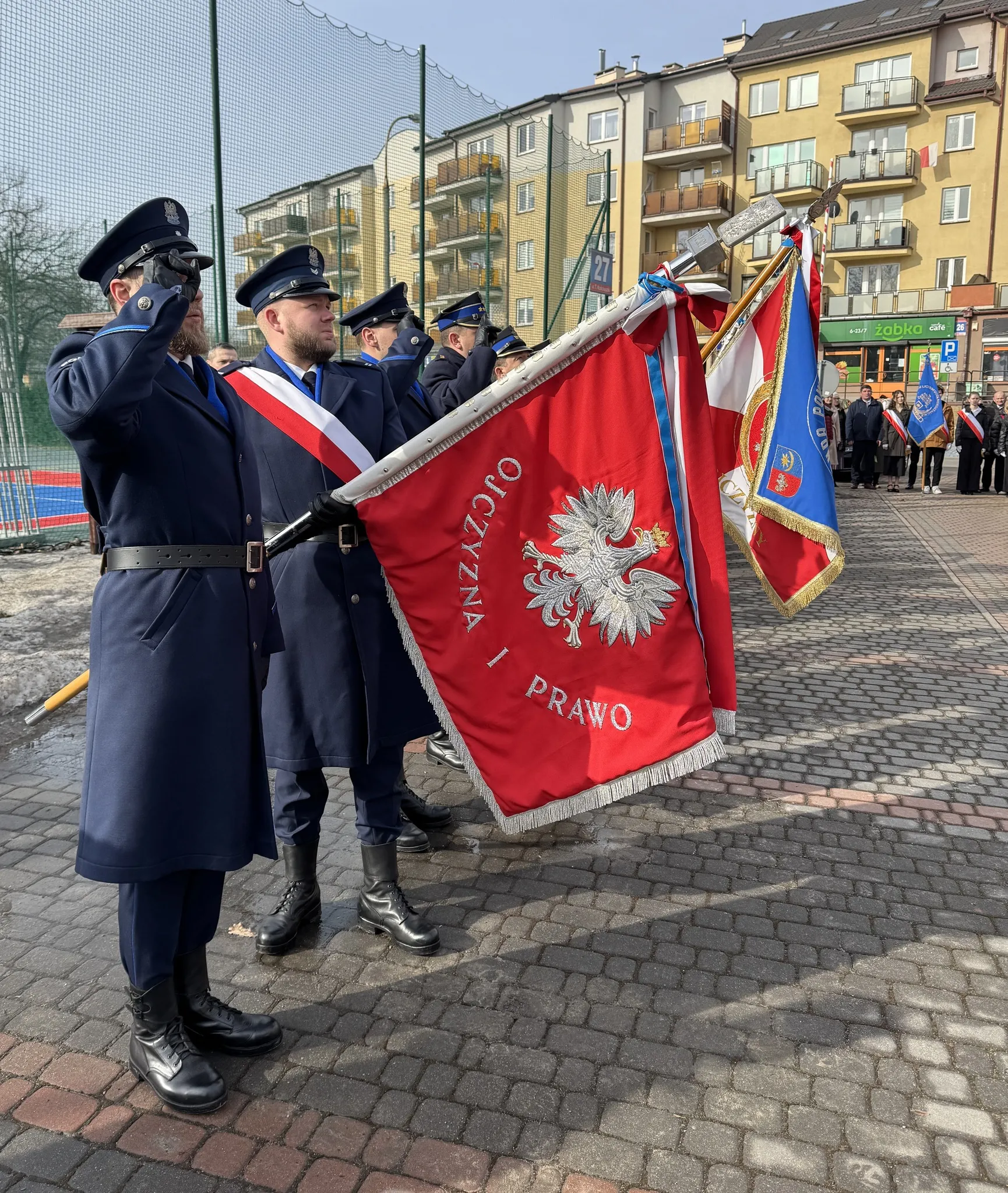 Ubrani w granatowe mundury mężczyźni stoją w szeregu i salutują, trzymając duży czerwony sztandar z białym orłem. W tle inne poczty sztandarowe i grupa ludzi na brukowanym placu przed blokami mieszkalnymi. Scena odbywa się na zewnątrz w ciągu dnia.
