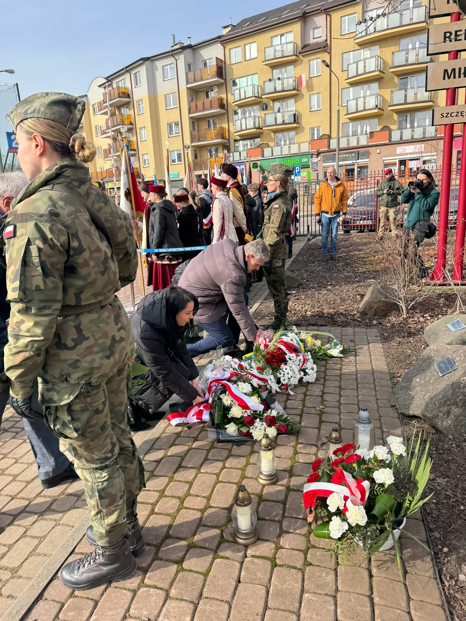 Dzień, zewnętrzne miejsce pamięci przy chodniku. Ludzie klękają i układają wieńce z biało-czerwonymi wstęgami oraz zapalają znicze. W tle stoją ubrani w mundury żołnierze i grupa osób z flagami przed blokami mieszkalnymi.