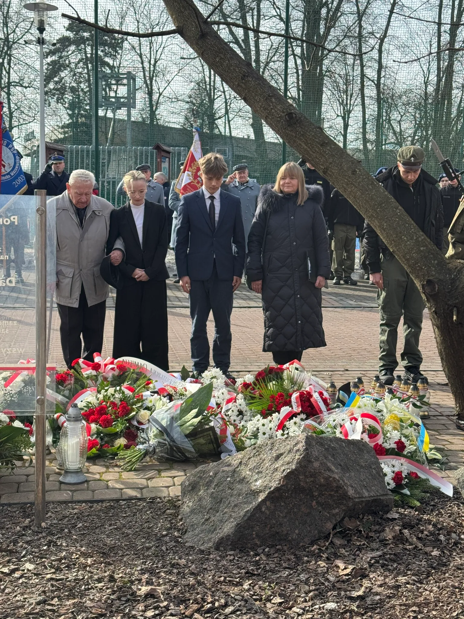 Grupa pięciu osób stoi przy ziemi z ułożonymi wieńcami i zniczami. Po prawej stoi mężczyzna w wojskowym mundurze, w tle widoczne flagi i kilka umundurowanych osób. Przed nimi na ziemi leży duży kamień i gałąź drzewa przebiega skośnie przez obraz.