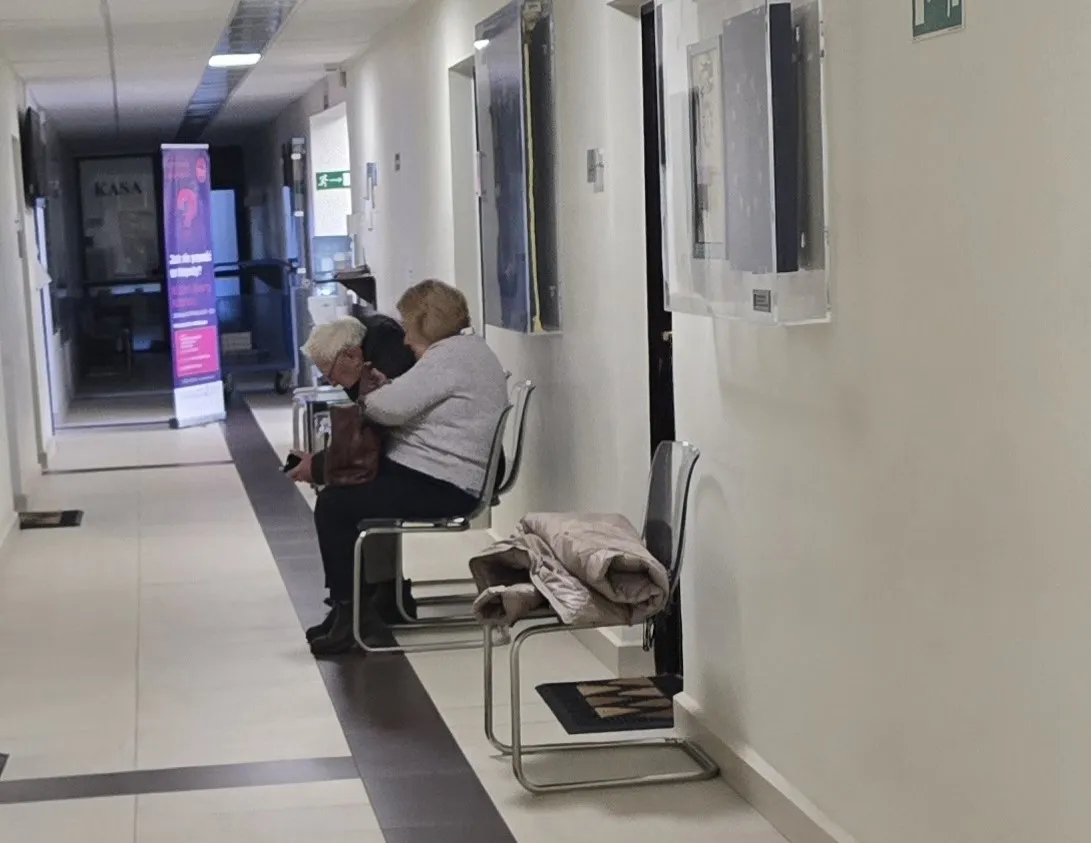 Korytarz z białymi ścianami i jasną podłogą z ciemnym paskiem na środku. Po prawej stronie trzy puste krzesła metalowe z czarnymi siedziskami, na jednym z nich złożony jasny płaszcz. Na dalszym planie dwie osoby siedzące na krzesłach, jedna starsza z siwymi włosami, druga młodsza, pochylone przed siebie. W tle widać drzwi i stojak z pionowym, kolorowym banerem informacyjnym.
