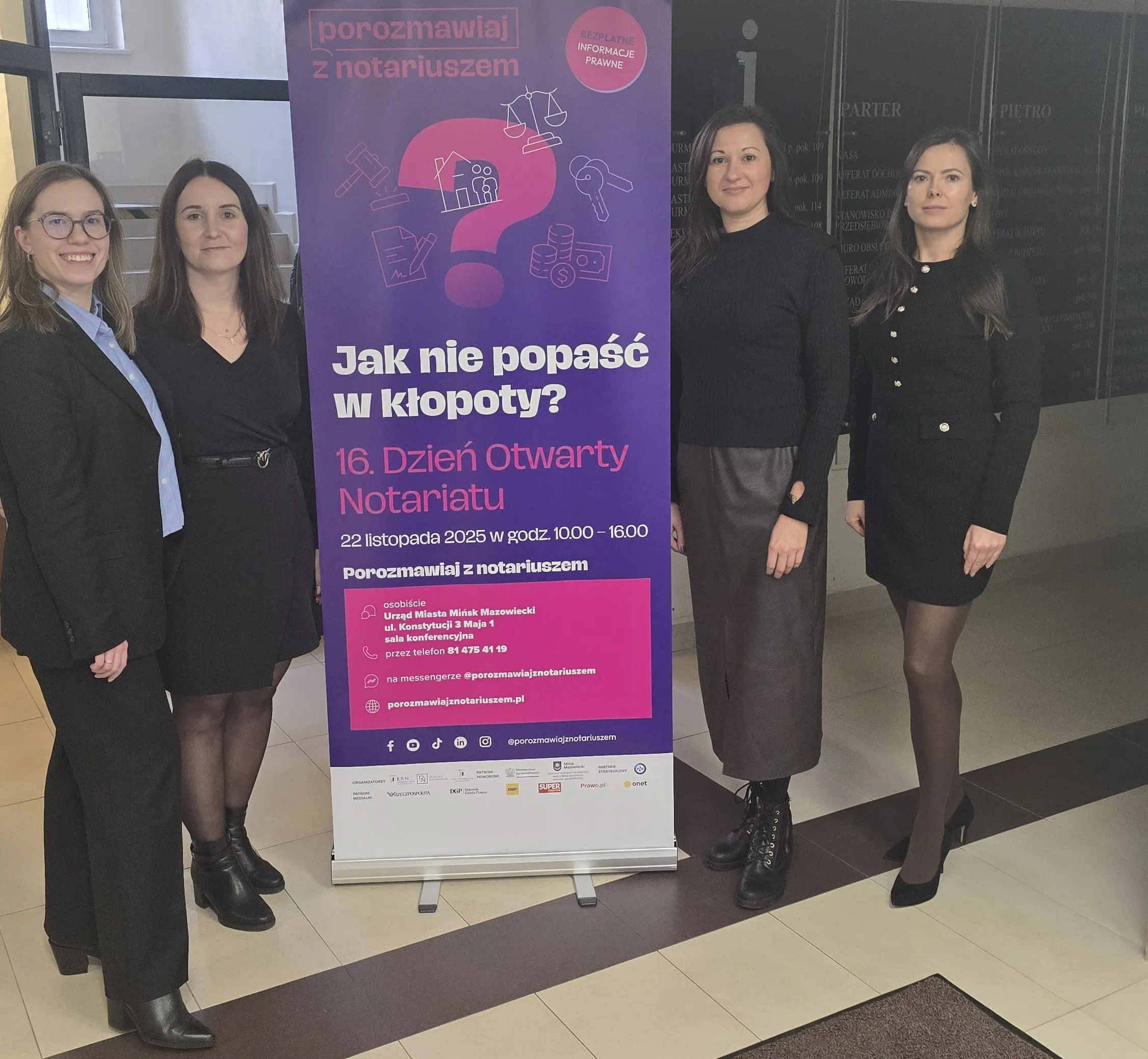 Cztery kobiety stoją obok pionowego baneru informacyjnego w jasnym wnętrzu. Baner ma fioletowe tło z różowymi i białymi napisami dotyczącymi Dnia Otwartego Notariatu. Kobiety są ubrane w eleganckie, ciemne ubrania.