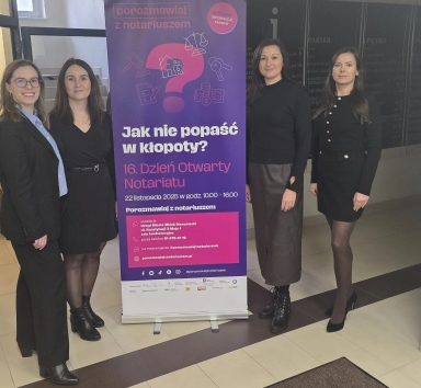 Cztery kobiety stoją obok pionowego baneru informacyjnego w jasnym wnętrzu. Baner ma fioletowe tło z różowymi i białymi napisami dotyczącymi Dnia Otwartego Notariatu. Kobiety są ubrane w eleganckie, ciemne ubrania.