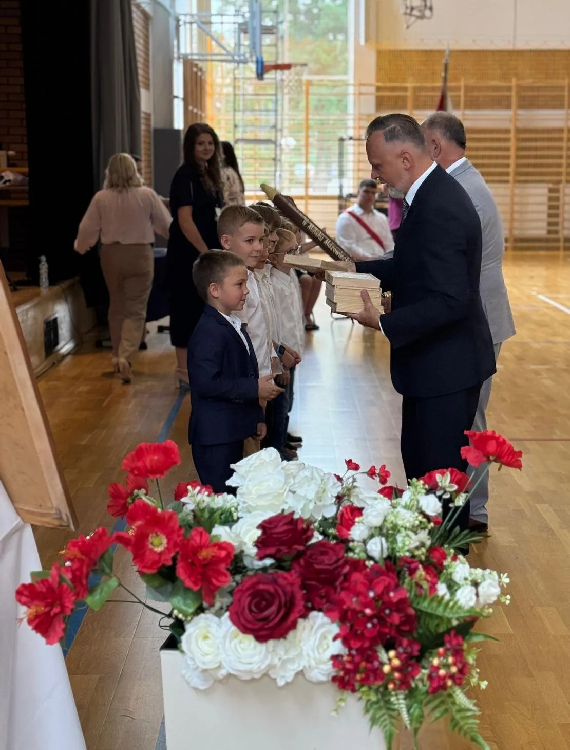 Mężczyzna w garniturze wręcza drewniane tabliczki grupie stojących chłopców w sali gimnastycznej, z przodu znajduje się bukiet czerwonych i białych kwiatów.