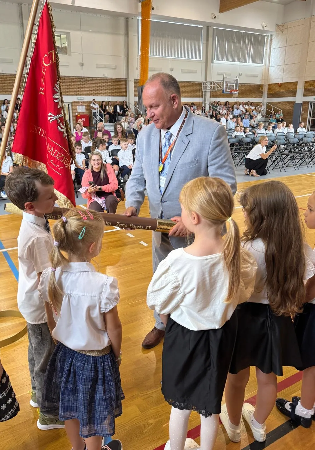 Mężczyzna w garniturze przekazuje dużą pałeczkę czterem dzieciom w hali sportowej podczas szkolnej uroczystości z obecnymi widzami na trybunach.