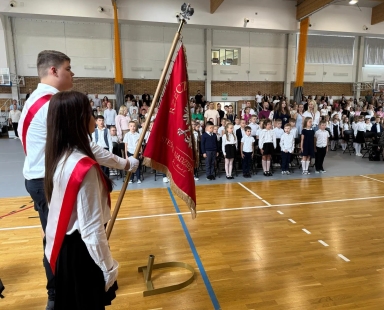 Dwóch uczniów w białych koszulach z czerwonymi szarfami trzymających sztandar szkolny stoi na sali gimnastycznej, a przed nimi stoi grupa dzieci i dorosłych, wszyscy ubrani formalnie.