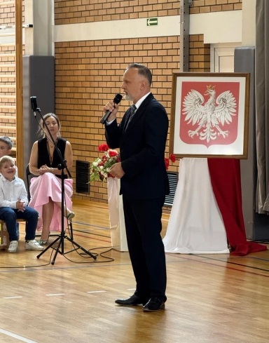 Mężczyzna w garniturze przemawia do publiczności w sali gimnastycznej, obok niego stoi godło Polski na tle biało-czerwonej tkaniny, a w tle siedzi kobieta i dzieci.