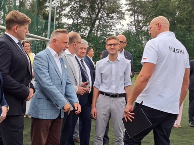 Grupa około ośmiu osób stoi na zewnątrz na boisku piłkarskim w ciągu dnia. Na środku stoi młodszy mężczyzna w paskowanej koszuli i okularach. Inni mężczyźni noszą marynarki i koszule. Po prawej stoi mężczyzna w białej koszulce z napisem POLSKA na plecach, w tle widać bramkę i drzewa.