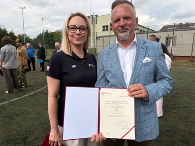 Dwie osoby stoją na boisku na zewnątrz. Kobieta w czarnej koszulce z krótkim rękawem i okularami stoi po lewej, mężczyzna w jasnoniebieskiej marynarce w kratę trzyma otwarty dokument z prawej. W tle ogrodzenie boiska i kilka osób rozmawiających.