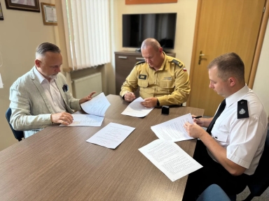 Oświetlone pomieszczenie. Trzech mężczyzn siedzi przy stole, przeglądając i podpisując dokumenty, z których jeden ma mundur służbowy straży pożarnej.