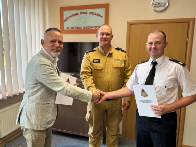 Trzech mężczyzn, w tym dwóch w mundurach Państwowej Straży Pożarnej, ściska sobie ręce i trzyma dokumenty w oświetlonym biurze.