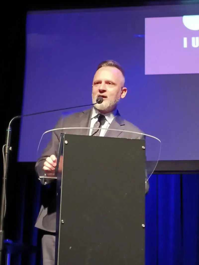 Mężczyzna w garniturze stoi za mównicą i przemawia do mikrofonu na tle niebieskiego ekranu.