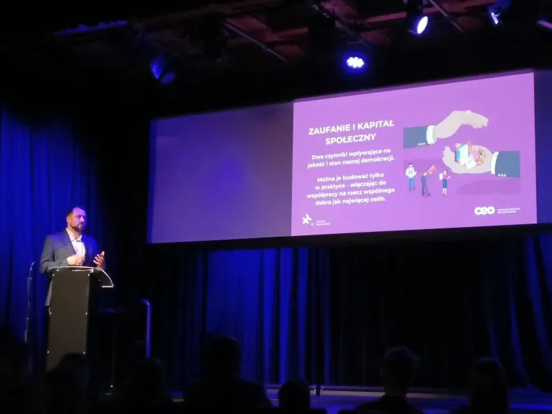 Mężczyzna przemawia przy mównicy na scenie przed ekranem z prezentacją zatytułowaną „Zaufanie i kapitał społeczny”.