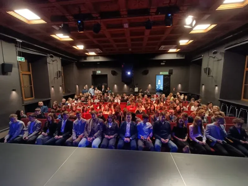 W sali teatralnej siedzi kilkadziesiąt osób skierowanych w stronę sceny.