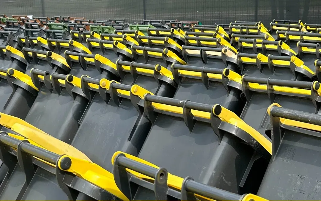 Rzędy szarych pojemników na odpady z żółtymi uchwytami ustawione na zewnątrz. 