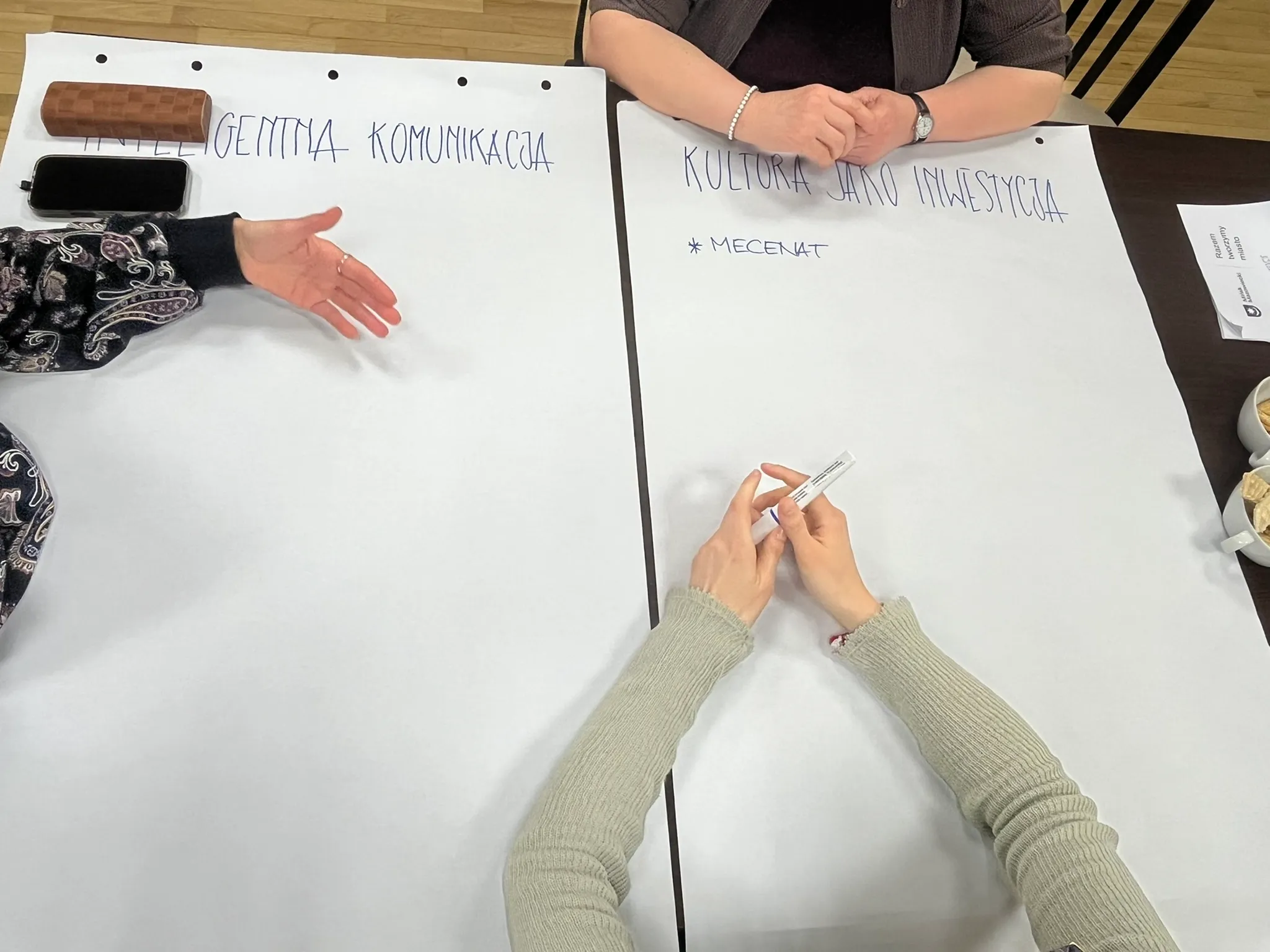 Dwie duże białe kartki rozłożone na ciemnym stole. Trzy pary rąk przy kartkach: jedna para po lewej gestykuluje, druga u góry ma złożone dłonie z bransoletką, trzecia u dołu trzyma marker. Na stole leży telefon i etui. Po prawej dwie filiżanki z przekąskami. U góry widoczna drewniana podłoga.