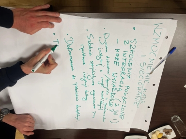 Dwie ręce piszą zielonym markerem na dużej białej kartce leżącej na ciemnym drewnianym stole. Na kartce są pionowo zapisane zielone notatki po polsku. Obok kartki leży niebieski długopis i talerzyki z przekąskami. Kartka ma trzy dziurki.